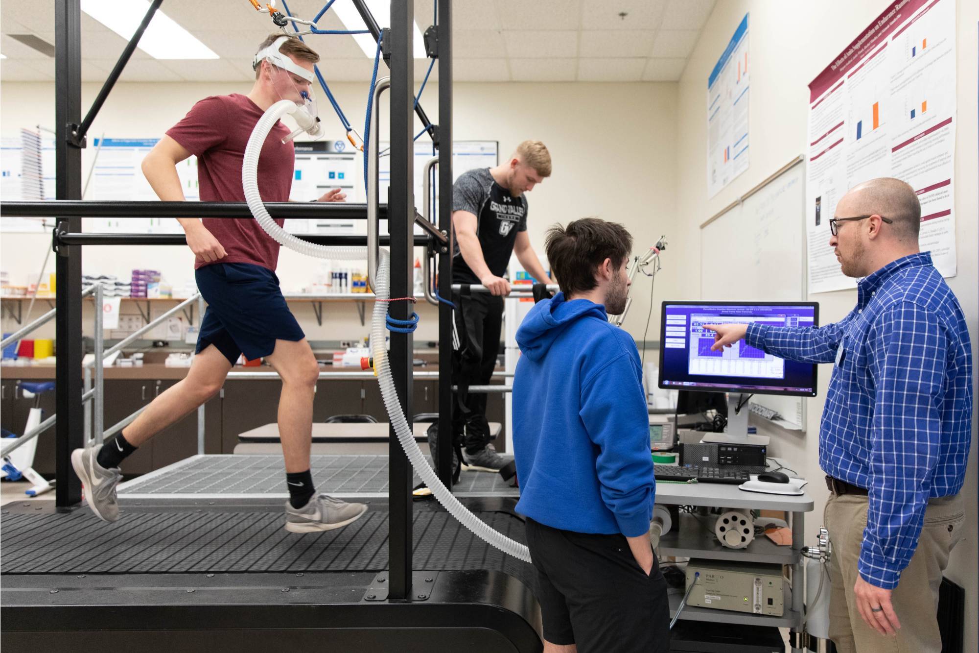 Faculty working with students performing VO2max test. One student is running on the treadmill with O2 testing equipment. Students with faculty reviewing computer screen and supervising.